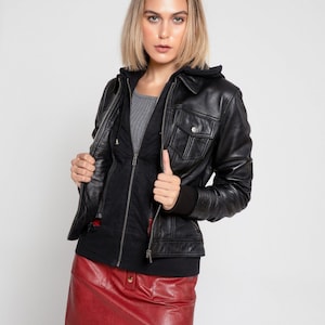 May include: A black leather jacket with a collared neckline and two chest pockets. The jacket is worn over a black hooded top and a gray shirt. A red leather mini-skirt completes the outfit.