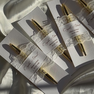 Op de afbeelding: Verschillende gouden pennen worden gepresenteerd op witte kaarten, elk versierd met een delicate kanten lint. Het lint bevat de tekst "Ceyda & Umut" en de datum "25/05/2026". De pennen hebben een glanzende gouden afwerking.