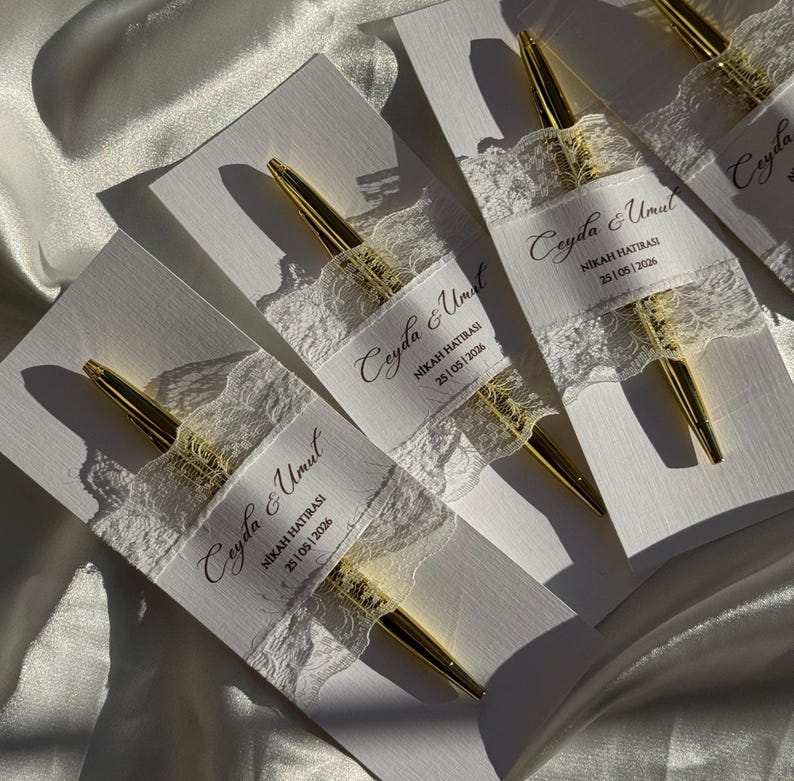 Op de afbeelding: Verschillende gouden pennen worden gepresenteerd op witte kaarten, elk versierd met een delicate kanten lint. Het lint bevat de tekst "Ceyda & Umut" en de datum "25/05/2026". De pennen hebben een glanzende gouden afwerking.