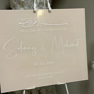Op de afbeelding: Een beige acrylen trouwteken met witte letters. Het bord bevat Arabische tekst, de woorden "WELCOME TO OUR BEGINNING", namen en de datum "10/02/2024". Onderaan staat een citaat uit de Koran.