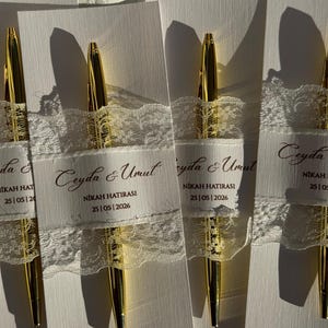Op de afbeelding: Vier gouden pennen worden getoond, elk versierd met een wit lint en kant. De linten dragen de tekst "Ceyda & Umut" en "NIKAH HATIRASI 25|05|2026". De pennen zijn geplaatst op een wit oppervlak, wat een strakke en elegante uitstraling cre&euml;ert.