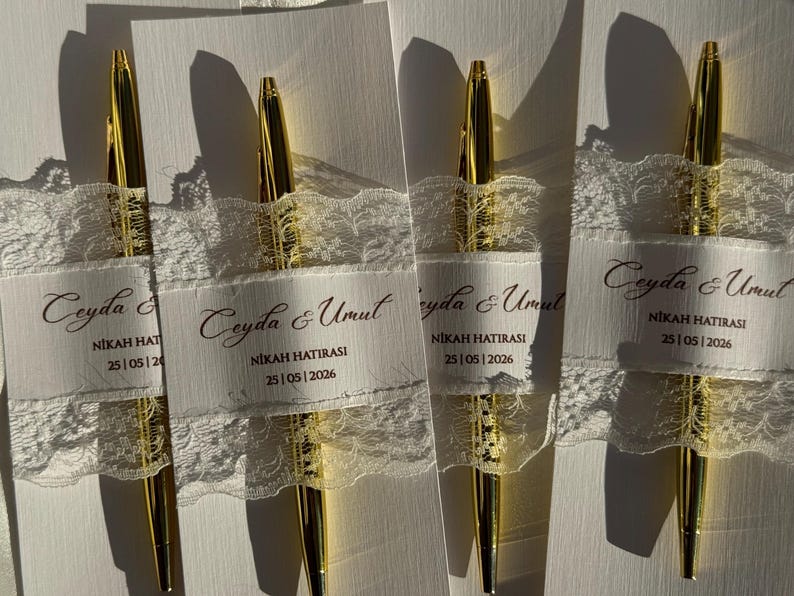 Op de afbeelding: Vier gouden pennen worden getoond, elk versierd met een wit lint en kant. De linten dragen de tekst "Ceyda & Umut" en "NIKAH HATIRASI 25|05|2026". De pennen zijn geplaatst op een wit oppervlak, wat een strakke en elegante uitstraling cre&euml;ert.