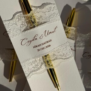 Op de afbeelding: Witte kaart met gouden pen, versierd met wit kant en tekst. De pen is goudkleurig met een glanzende afwerking. De kaart heeft de tekst "Ceyda & Umut", "NİKAH HATIRASI" en de datum "25 | 05 | 2026".