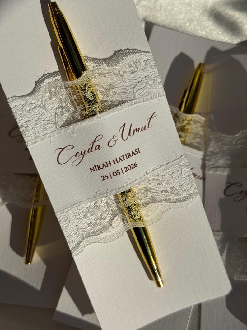 Op de afbeelding: Witte kaart met gouden pen, versierd met wit kant en tekst. De pen is goudkleurig met een glanzende afwerking. De kaart heeft de tekst "Ceyda & Umut", "NİKAH HATIRASI" en de datum "25 | 05 | 2026".