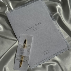 Op de afbeelding: Een witte, getextureerde papieren map en een bijpassende rechthoekige kaart met een gouden pen. De map is voorzien van kanten randen en de tekst "Zamir & Nabila", "NEKHA KHAT", "13.09.2026" en Arabisch schrift. De kaart toont ook de namen en datum.