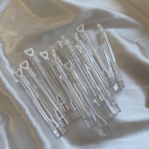 Op de afbeelding: Een verzameling doorzichtige plastic bellenblaasstokjes, elk voorzien van een witte, hartvormige handgreep en dop. Deze slanke, cilindervormige houders zijn ontworpen voor het blazen van bellen en liggen in een groepje.