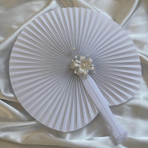 Op de afbeelding: Een ronde witte handwaaier met een geplooid papierontwerp en een wit plastic handvat. Het midden van de waaier is versierd met een zilverkleurige metalen bloem met kleine witte kunstparels en dunne draadstengels die naar buiten steken.