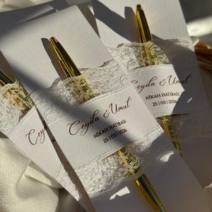 Op de afbeelding: Gouden pennen met witte kaarten, versierd met kant en de tekst "Ceyda & Umut NIKAH HATIRASI 25|05|2026". De pennen liggen in een mandje, badend in natuurlijk licht.