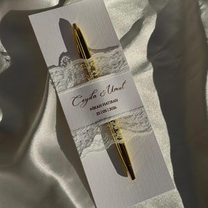 Op de afbeelding: Een gouden pen ligt op een witte kaart, vastgehouden door een wit kanten lint. De kaart heeft gouden tekst: "Ceyda & Umut, NIKAH HATIRASI, 25|05|2026". De achtergrond is een witte, satijnachtige stof.