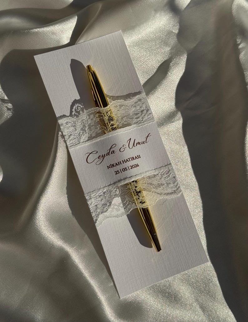 Op de afbeelding: Een gouden pen ligt op een witte kaart, vastgehouden door een wit kanten lint. De kaart heeft gouden tekst: "Ceyda & Umut, NIKAH HATIRASI, 25|05|2026". De achtergrond is een witte, satijnachtige stof.