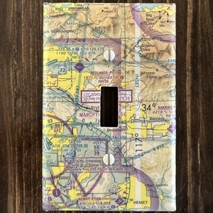 May include: A light switch cover featuring a colorful map design with yellow, purple, and green hues. The cover has a rectangular shape with a single switch opening and two screw holes. The background is a dark brown wooden surface.