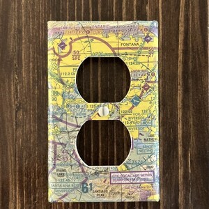 May include: A decorative light switch cover featuring a detailed aviation sectional chart design. The cover has two circular openings and a screw, set against a dark wood background. The chart includes place names and numbers.