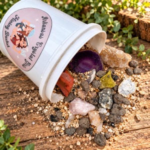 May include: A white plastic bucket labeled Arkansas Crystal Gus Mining Bucket, tipped over to reveal a collection of raw minerals including a purple agate slice, a pink rose quartz, a red stone, and various small grey, yellow, and clear rocks.