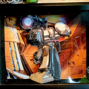 Może przedstawiać: Ramka z 3D wyskakującą grafiką przedstawiającą srebrnego robota z dużym pistoletem, stojącego na tle żółtym i pomarańczowym.