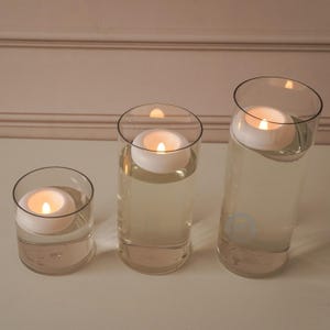 Pode incluir: Tr&ecirc;s cilindros de vidro transparente de alturas variadas, cada um preenchido com &aacute;gua e uma vela branca flutuante. As velas est&atilde;o acesas, emitindo um brilho quente. A disposi&ccedil;&atilde;o est&aacute; sobre uma superf&iacute;cie branca, com um fundo neutro.