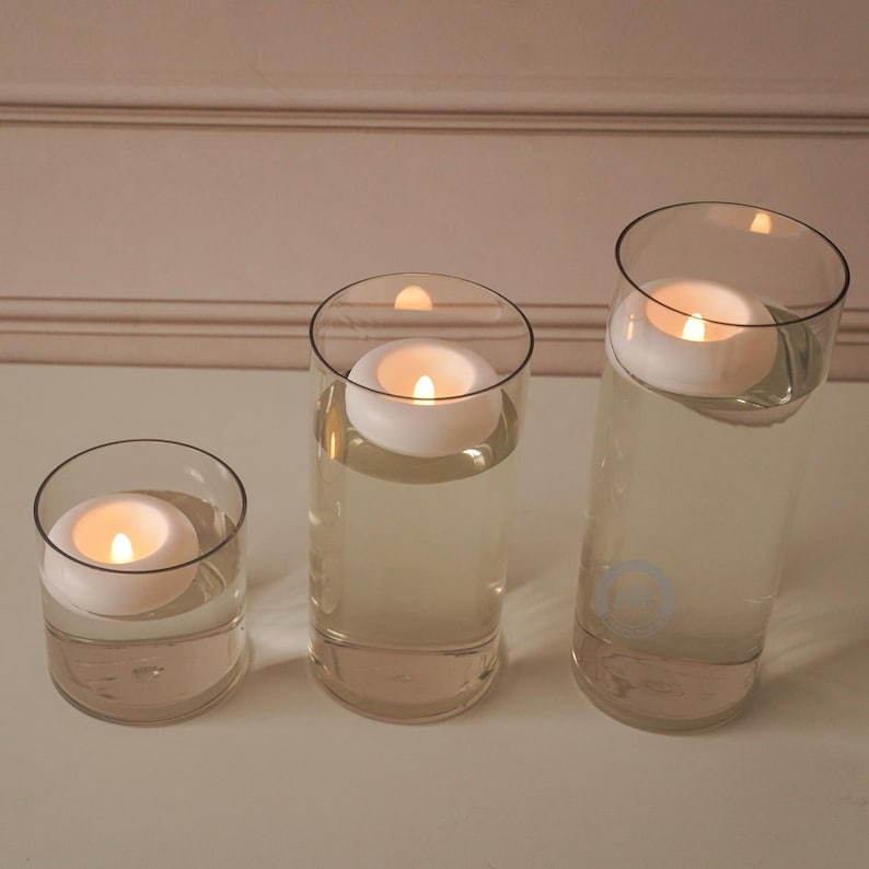 Pode incluir: Tr&ecirc;s cilindros de vidro transparente de alturas variadas, cada um preenchido com &aacute;gua e uma vela branca flutuante. As velas est&atilde;o acesas, emitindo um brilho quente. A disposi&ccedil;&atilde;o est&aacute; sobre uma superf&iacute;cie branca, com um fundo neutro.