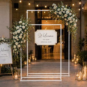 Puede incluir: Decoración de ceremonia de boda con arreglos florales blancos y letreros que dicen "Bienvenido a nuestro día especial". La escena incluye un arco blanco, velas y luces de cadena.