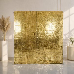 May include: A large, free-standing gold sequin backdrop. The backdrop is made of many small, reflective gold sequins, creating a shimmering effect. The backdrop is rectangular and stands upright, suitable for events and photo opportunities.