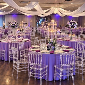 May include: A banquet hall featuring round tables covered in purple tablecloths, surrounded by clear acrylic chairs. Each table is set with white plates, glassware, and a tall centerpiece with candles and purple and white floral arrangements.