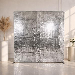 May include: A large, silver sequin backdrop stands upright, reflecting light. The backdrop is made of many small, square panels. The backdrop is designed for events and photo shoots. The backdrop is in a bright, well-lit room.