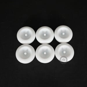 Pode incluir: Seis objetos circulares brancos com um ponto central elevado est&atilde;o dispostos em duas filas de tr&ecirc;s sobre um fundo preto. Os objetos parecem ser feitos de um material liso e mate.