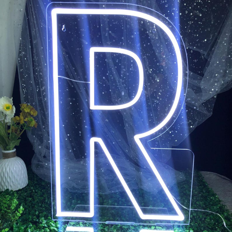 May include: A large, illuminated letter "R" in a neon blue glow. The letter is outlined in bright white light, creating a striking contrast against the dark background. The neon sign is a decorative piece, possibly for a room or event.