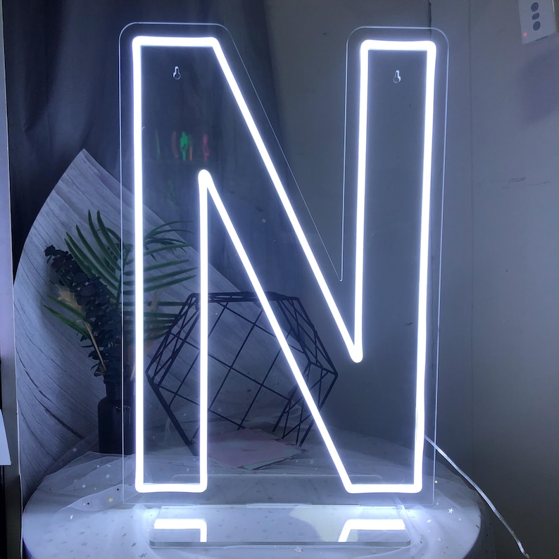 May include: A white neon light shaped like the letter "N". The neon sign is mounted on a clear acrylic base. The illuminated letter is bright against a neutral background. The sign is plugged in and ready to use.