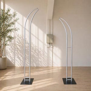 Könnte beinhalten: Zwei silberne Metall-Bogenrahmen mit gewölbter Oberseite und rechteckiger Basis. Die Rahmen stehen auf einem hellbraunen Holzboden. Der Hintergrund ist eine hellbeige Wand.