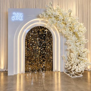 May include: A white arched backdrop featuring a glowing neon sign that reads MIS 15 QUINCE. The arch interior is filled with shimmering gold square sequins, and a large, dense arrangement of white cherry blossom branches curves along the right side.