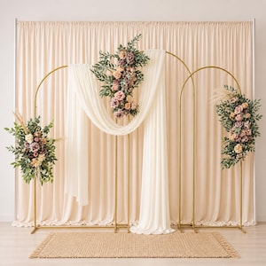 May include: A gold metal wedding arch with cream-colored fabric and floral arrangements. The arch is decorated with pink, cream, and green flowers and greenery. A beige rug is in front of the arch, and a cream-colored curtain is in the background.
