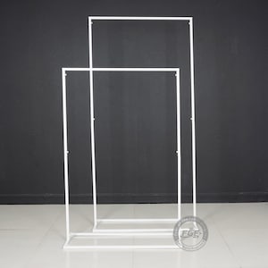 May include: Two white metal arch frames, one taller than the other, standing on a gray floor. The frames are made of thin metal tubing and are designed to be used as a backdrop for events or displays.