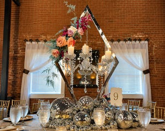 Pode incluir: Um centro de mesa com uma moldura prateada em forma de diamante que sustenta um candelabro de cristal com velas brancas, adornado com flores cor-de-rosa e laranja. A base é composta por várias bolas de discoteca prateadas e um número 9.