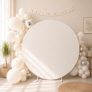 Op de afbeelding: Een witte, ronde achtergrond voor een feest, ondersteund door een metalen frame. De achtergrond is omgeven door een ballonnenboog in wit en zilver, met een banner met de tekst "HAPPY BIRTHDAY". Een potplant en een kleine tafel met bloemen zijn ook zichtbaar.