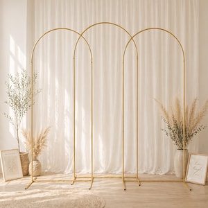 May include: Three gold metal arch wedding backdrops. The arches are arranged in a row, with a rectangular base and a curved top. The backdrop is set against a white curtain. Decorative plants and framed art pieces are on either side.