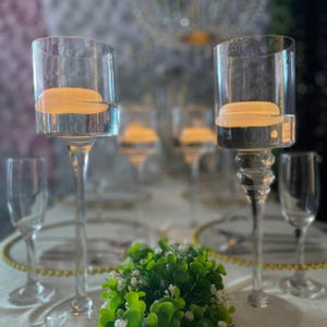 Pode incluir: Casti&ccedil;ais de vidro transparente com um topo cil&iacute;ndrico e uma haste longa e elegante. Cada casti&ccedil;al cont&eacute;m uma vela flutuante. A mesa inclui ta&ccedil;as de champanhe e vegeta&ccedil;&atilde;o decorativa, sugerindo um evento formal ou um casamento.
