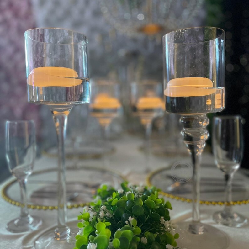 Pode incluir: Casti&ccedil;ais de vidro transparente com um topo cil&iacute;ndrico e uma haste longa e elegante. Cada casti&ccedil;al cont&eacute;m uma vela flutuante. A mesa inclui ta&ccedil;as de champanhe e vegeta&ccedil;&atilde;o decorativa, sugerindo um evento formal ou um casamento.