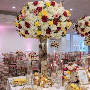 May include: A tall, mirrored centerpiece stand topped with a large, spherical floral arrangement of white, yellow, pink, and deep red roses. The base features a gold-framed sign with the letter B, surrounded by smaller floral bouquets and candles.