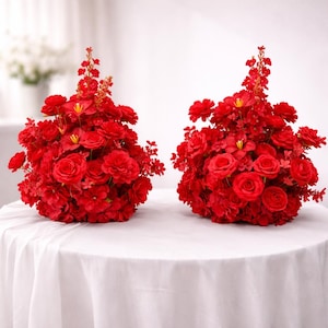 Puede incluir: Dos arreglos florales rojos vibrantes, cada uno con un denso grupo de rosas y otras flores. Los arreglos están sobre un mantel blanco, creando un contraste llamativo. Las flores son de un rojo intenso y rico, perfectas para centros de mesa o exhibiciones decorativas.