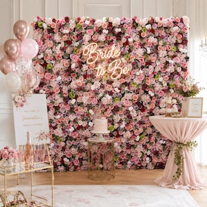 May include: A large floral wall backdrop featuring a dense arrangement of pink, red, and white roses with a neon sign reading Bride to Be. Includes a balloon cluster, a welcome sign for a bridal shower, a cake stand, and a draped table.