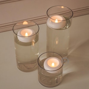 May include: Three clear glass candle holders with floating white candles. The candles are lit, casting a warm glow. The holders are filled with a clear liquid, creating a serene and elegant display. Perfect for home decor or special events.