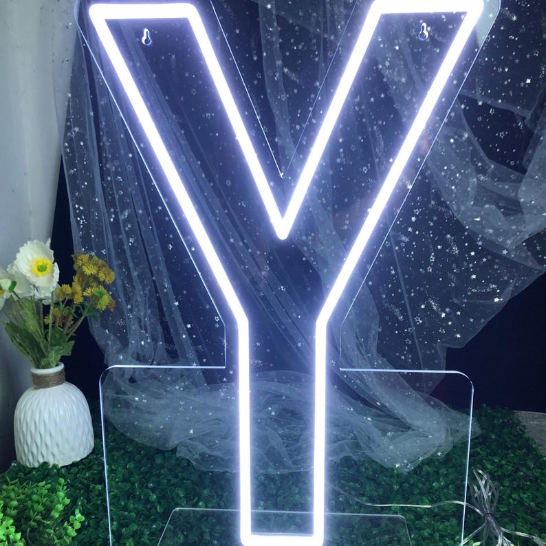 May include: A glowing, neon-style letter "Y" in bright white, set against a backdrop of a sheer, starry fabric. The letter is mounted on a clear base, with a vase of flowers to the left. The overall effect is modern and decorative.