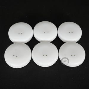 Pode incluir: Seis bot&otilde;es brancos e redondos, cada um com dois pequenos orif&iacute;cios, dispostos em duas filas sobre um fundo preto. Os bot&otilde;es t&ecirc;m uma superf&iacute;cie lisa e ligeiramente abaulada. Estes bot&otilde;es s&atilde;o provavelmente para costura ou artesanato.