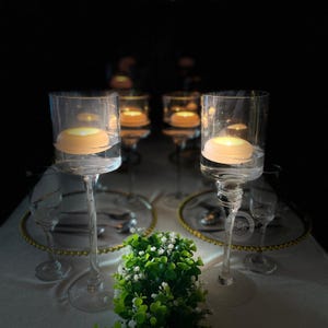 May include: Clear glass candle holders with floating candles on a white tablecloth. The candle holders have a tall, slender stem and a wide, cylindrical top. The table setting includes plates, silverware, and small green and white floral arrangements.