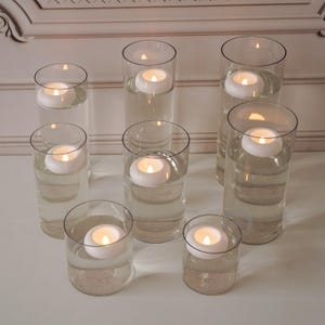 Pode incluir: Uma cole&ccedil;&atilde;o de casti&ccedil;ais de vidro transparente cheios de &aacute;gua, cada um com uma vela branca flutuante com uma chama acesa. As velas est&atilde;o dispostas sobre uma superf&iacute;cie branca, criando um brilho suave e quente. O fundo apresenta uma parede decorativa.