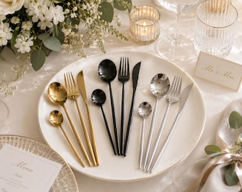 May include: A set of three flatware groupings arranged on a white plate, featuring gold, black, and silver finishes. Each set includes a dinner spoon, a dinner fork, a dinner knife, and a small teaspoon, all with slender handles and polished surfaces.