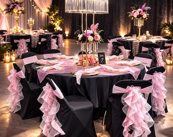 May include: A round table with a black tablecloth, pink runner, and chairs featuring pink ruffled sashes. A tall gold rectangular centerpiece with hanging crystals and a floral arrangement sits in the center, with a small black table number sign.