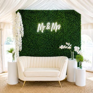 May include: A wedding reception setup featuring a cream-colored loveseat with gold legs, set against a green hedge wall with a neon "Mr & Mrs" sign. White floral arrangements and cylindrical pedestals complete the elegant decor.