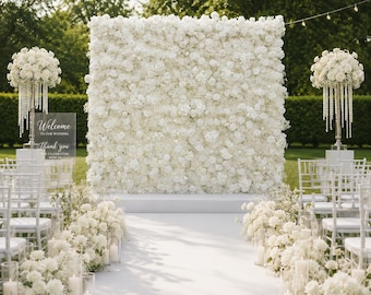 May include: A large square white floral backdrop wall featuring dense clusters of white roses and baby's breath. Two tall silver stands hold white floral arrangements with hanging crystal beads. A clear sign reads Welcome to our wedding.