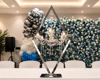 May include: A silver-toned metal diamond-shaped frame stand featuring a central crystal chandelier with multiple arms and hanging glass droplets. The structure is positioned on a flat base, designed to hold the chandelier securely in place.