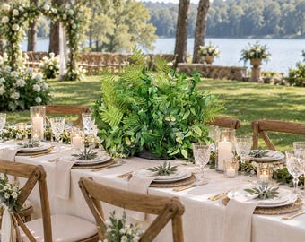 May include: A long dining table set for an outdoor event, featuring a beige linen tablecloth, white plates, clear crystal glassware, and white pillar candles. A large, lush green fern and foliage centerpiece sits in the center of the table.